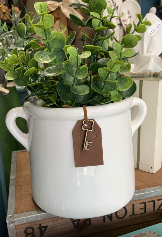Reverse side of a white ceramic flower pot with green plant and brown tag and key on a wooden surface