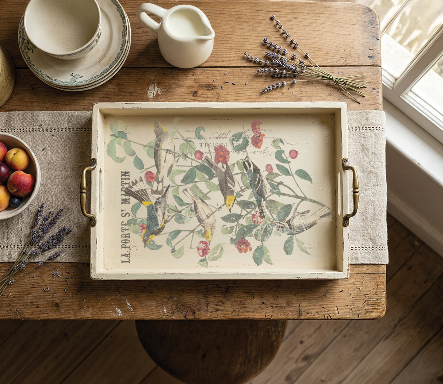 A rectangular serving tray with brass handles, featuring a vintage-style botanical illustration of birds and cherries with the text 'LA PORTS S. MARTIN', displayed on a rustic wooden table with a placemat, fruit bowl, lavender, and cream-colored ceramic items. From Iron Orchid Designs
