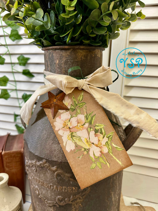 Decorative rusty milk churn with greenery, ribbon, and floral tag on a neutral background