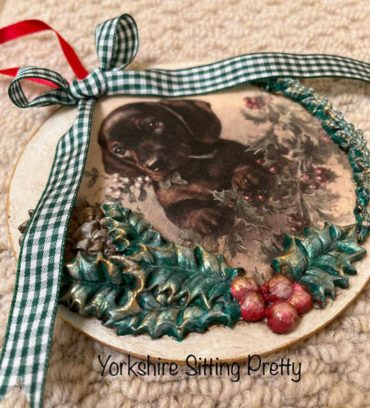 Flat circular Christmas bauble decorated with an image of a brown puppy with holly leaves and berries and a green gingham bow