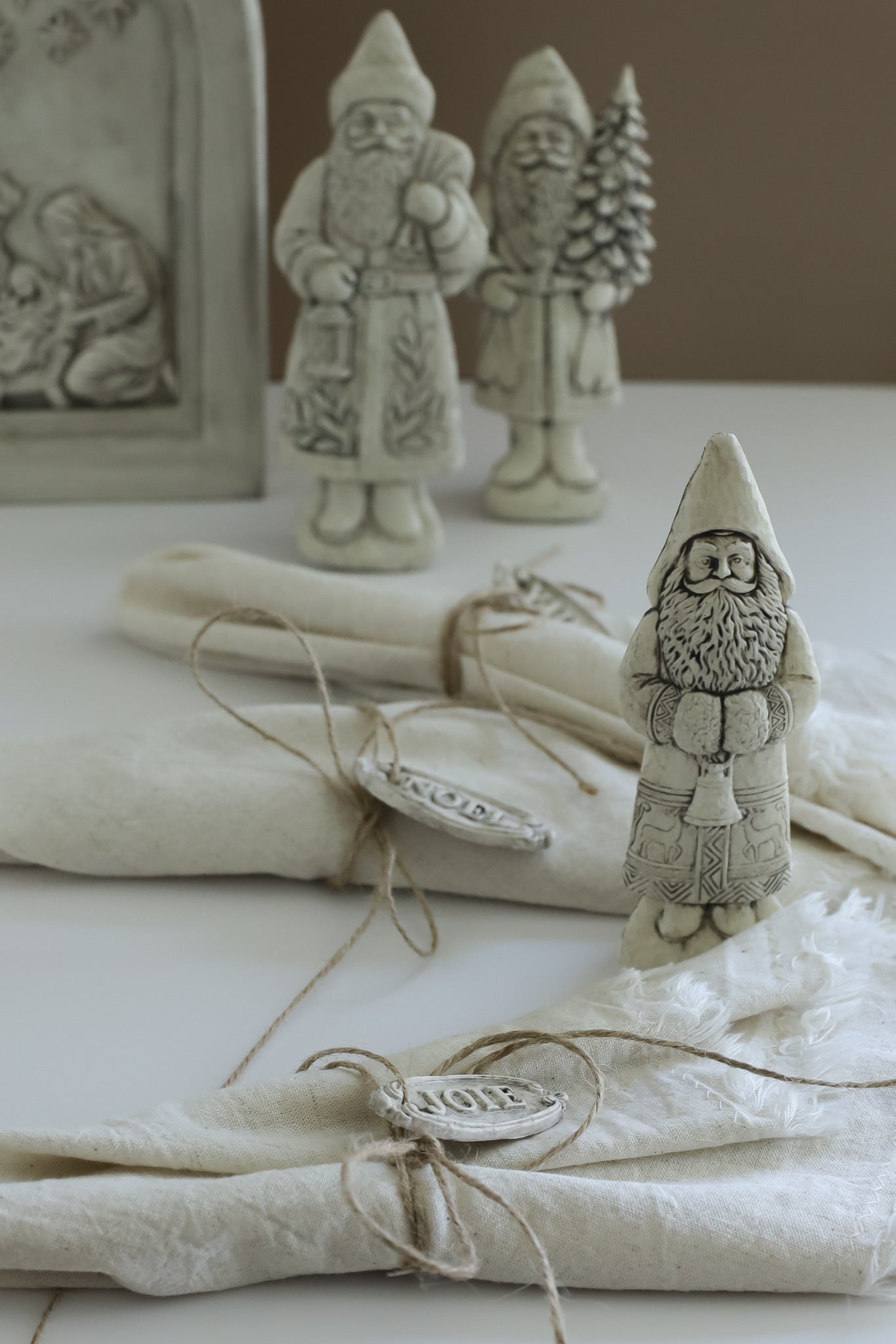 A styled tabletop scene with cream-colored fabric napkins tied with twine and small decorative Santa Claus figurines, including one in the foreground and two larger ones in the background.From Iron Orchid Designs.