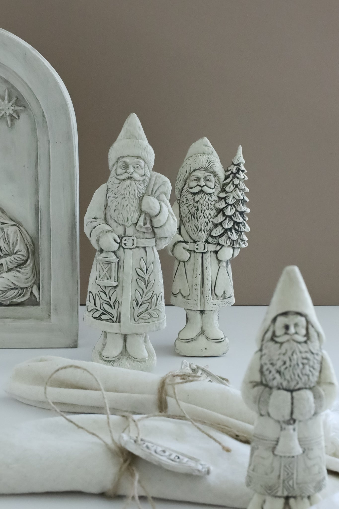 Three rustic white ceramic Santa Claus figurines with detailed carvings, displayed on a white surface with a neutral beige background. Belsnickel from Iron Orchid Designs.