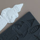 A dark gray silicone mold with a Santa Claus design shown next to a white molded Santa Claus product on a solid taupe background. Belsnickel From Iron Orchid Designs.