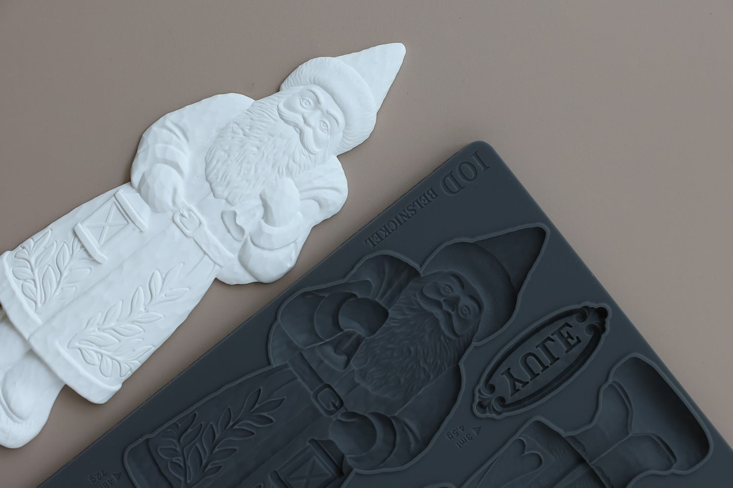 A dark gray silicone mold with a Santa Claus design shown next to a white molded Santa Claus product on a solid taupe background. Belsnickel From Iron Orchid Designs.