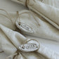 Three folded cream linen napkins tied with twine and decorated with oval white metal tags engraved with 'JOIE', 'NOEL', and 'Yule', arranged on a white surface.l Besnickel from Iron Orchid Designs.