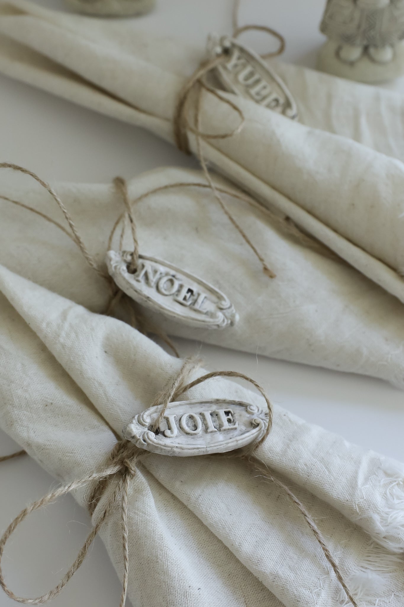 Three folded cream linen napkins tied with twine and decorated with oval white metal tags engraved with 'JOIE', 'NOEL', and 'Yule', arranged on a white surface.l Besnickel from Iron Orchid Designs.