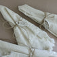 Three folded cream-colored linen napkins with frayed edges, tied with twine and decorated with silver metal nameplates spelling 'Joie', 'Yule',  arranged on a solid taupe background. Belsnickel From Iron Orchid Designs.