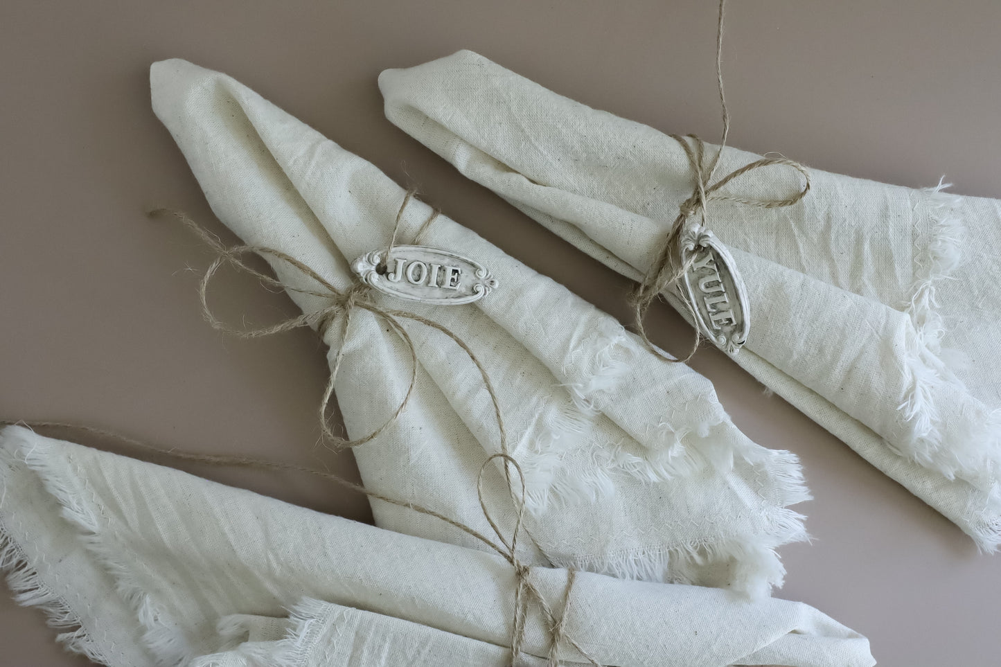 Three folded cream-colored linen napkins with frayed edges, tied with twine and decorated with silver metal nameplates spelling 'Joie', 'Yule',  arranged on a solid taupe background. Belsnickel From Iron Orchid Designs.
