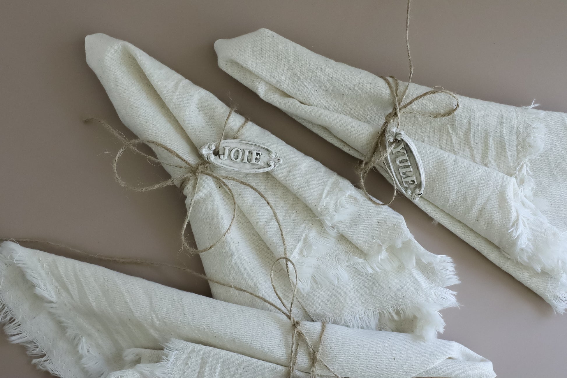 Three folded cream-colored linen napkins with frayed edges, tied with twine and decorated with silver metal nameplates spelling 'Joie', 'Yule',  arranged on a solid taupe background. Belsnickel From Iron Orchid Designs.