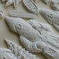 A close-up of several white carved Christmas ornaments and figurines arranged on a light beige surface, including a gnome, a Christmas tree, and a small sign reading "Old Colchester Road.". From Iron Orchid Designs.