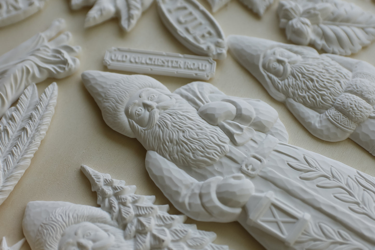 A close-up of several white carved Christmas ornaments and figurines arranged on a light beige surface, including a gnome, a Christmas tree, and a small sign reading "Old Colchester Road.". From Iron Orchid Designs.