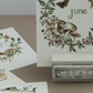 A decorative monthly calendar page holder displaying 'June' with a woodland bunny and floral wreath illustration, shown with other partial calendar pages and a small metal stand on a neutral tabletop. From Iron Orchid Designs