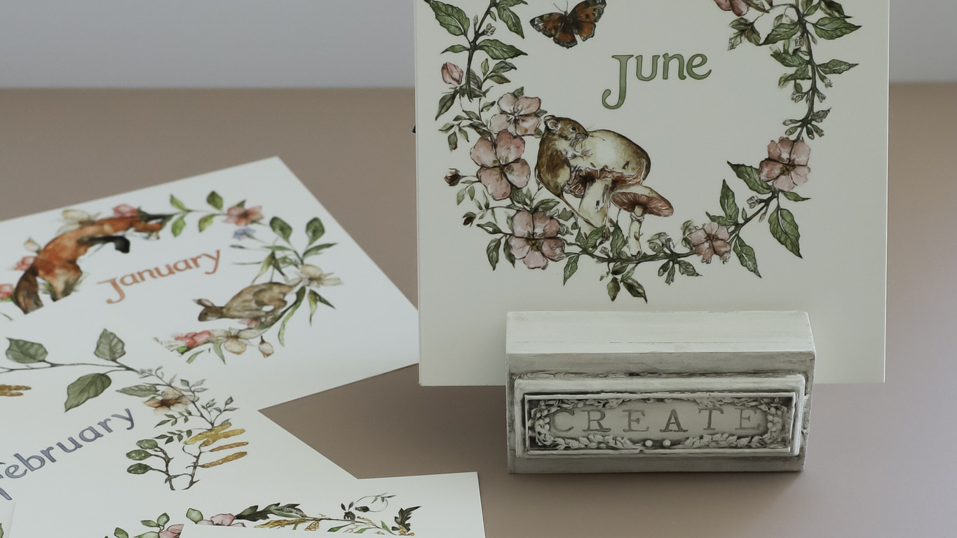 A decorative monthly calendar page holder displaying 'June' with a woodland bunny and floral wreath illustration, shown with other partial calendar pages and a small metal stand on a neutral tabletop. From Iron Orchid Designs