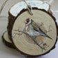A round wooden slice ornament with a hanging twine loop, featuring a painted goldfinch on a branch, with stacked similar slices in the background. From Iron Orchid Designs