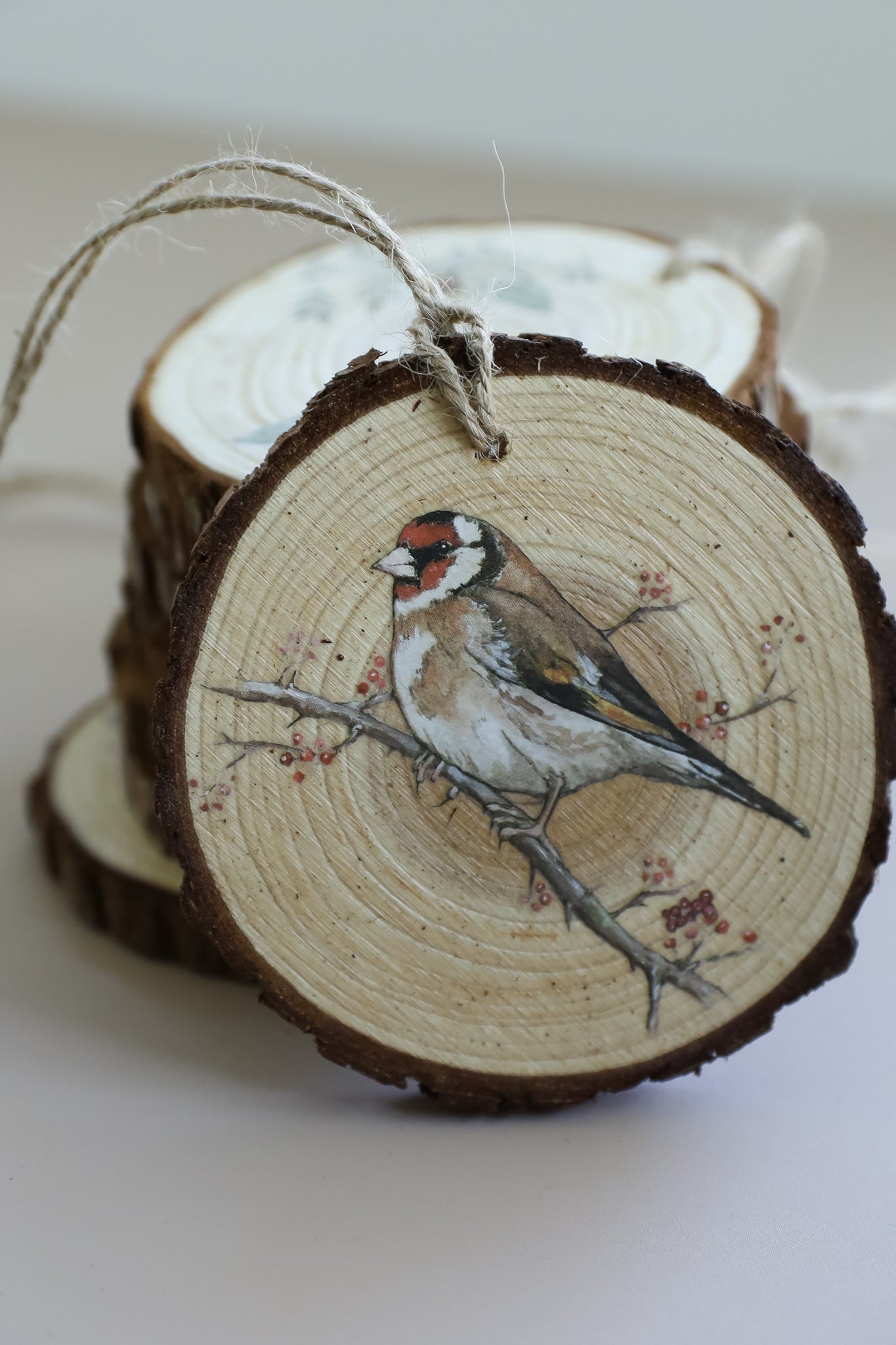A round wooden slice ornament with a hanging twine loop, featuring a painted goldfinch on a branch, with stacked similar slices in the background. From Iron Orchid Designs