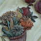 A close-up of a carved decorative wall piece showing a harvest basket filled with corn, pumpkins, gourds, and leaves, alongside pomegranates, on a light wood background. Cornucopias moulds. From Iron Orchid Designs.