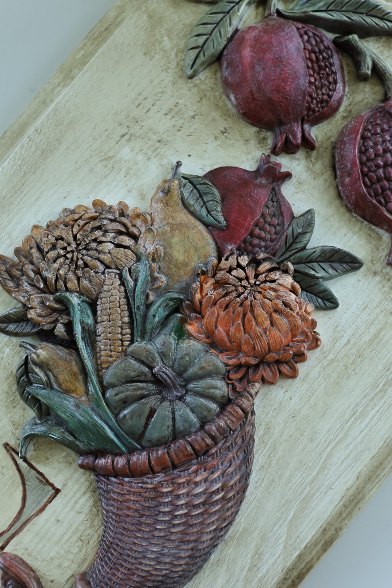 A close-up of a carved decorative wall piece showing a harvest basket filled with corn, pumpkins, gourds, and leaves, alongside pomegranates, on a light wood background. Cornucopias moulds. From Iron Orchid Designs.