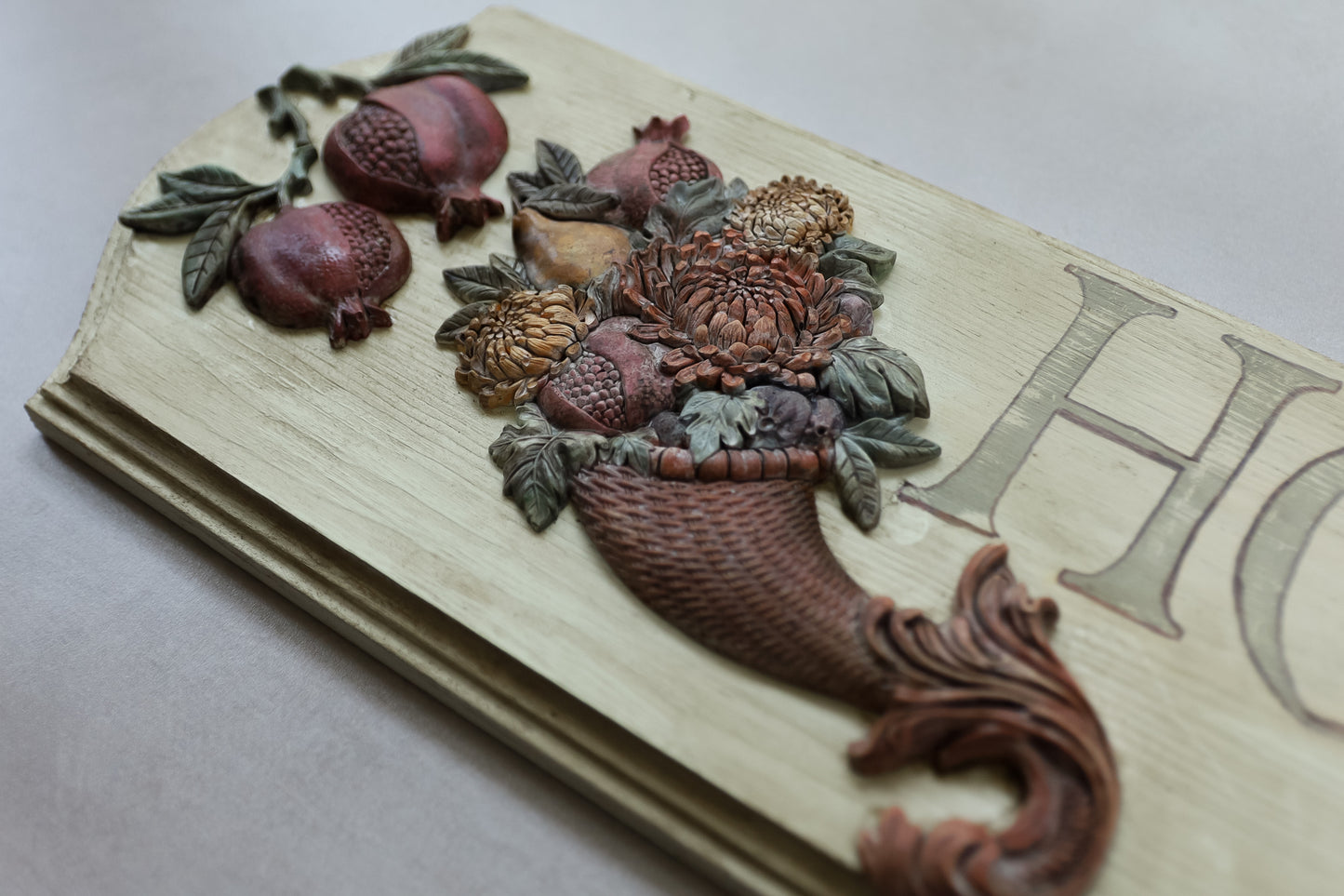 A close-up of a carved wooden decorative plaque featuring pomegranates, flowers, and a basket motif, with part of a large letter 'H' visible on the right side. Cornucopias mould. From Iron Orchid Designs.