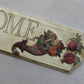 A decorative wooden sign with the word "HOME" in raised letters, featuring a carved cornucopia filled with fruits and vegetables, plus two pomegranates, on a light gray background. Cornucopias mould. From Iron Orchid Designs.