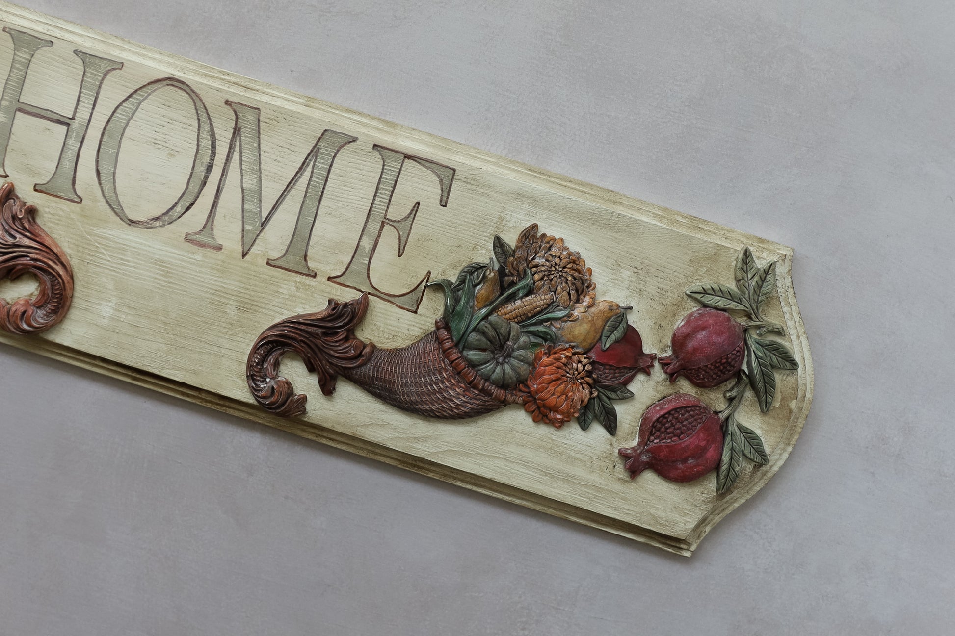 A decorative wooden sign with the word "HOME" in raised letters, featuring a carved cornucopia filled with fruits and vegetables, plus two pomegranates, on a light gray background. Cornucopias mould. From Iron Orchid Designs.
