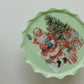 A scalloped-edge ceramic plate with a light mint green base, featuring a vintage illustration of Santa Claus, Christmas trees, and wrapped gifts on the top. From Iron Orchid Designs.