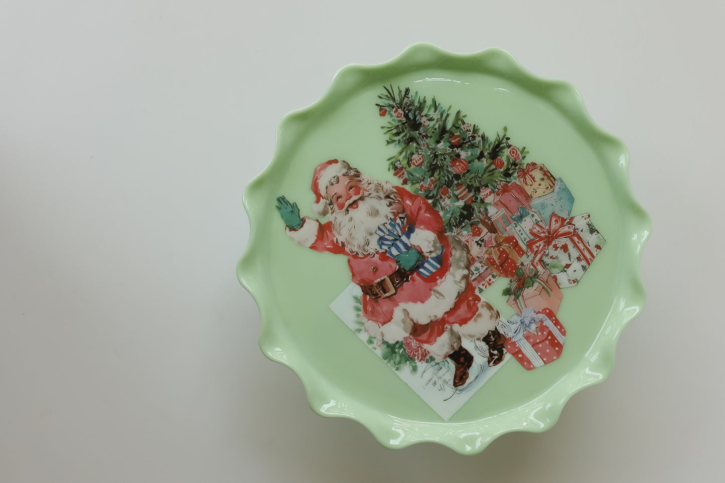 A scalloped-edge ceramic plate with a light mint green base, featuring a vintage illustration of Santa Claus, Christmas trees, and wrapped gifts on the top. From Iron Orchid Designs.