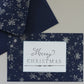 A navy blue Christmas greeting card with a white snowflake pattern and a white center label reading 'Merry Christmas,' shown with matching navy envelope pieces on a light neutral background. Flurry stamp From Iron Orchid Designs.