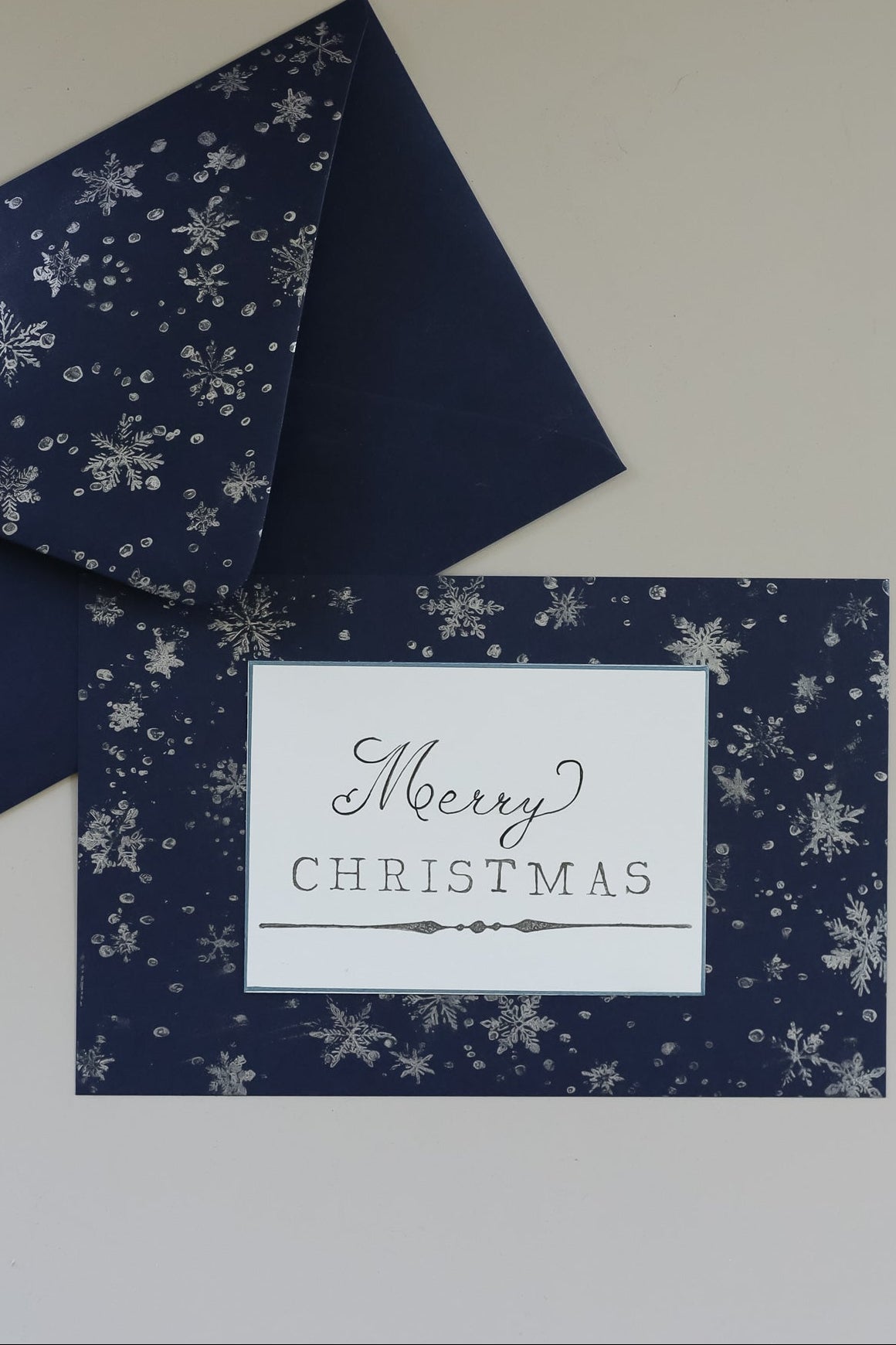 A navy blue Christmas greeting card with a white snowflake pattern and a white center label reading 'Merry Christmas,' shown with matching navy envelope pieces on a light neutral background. Flurry stamp From Iron Orchid Designs.