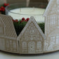 A close-up of a decorative holiday candle holder made from three gingerbread house cutouts in beige and white, surrounding a glass candle with white wax and red berries. Gingerbread Lane From Iron Orchid Designs.