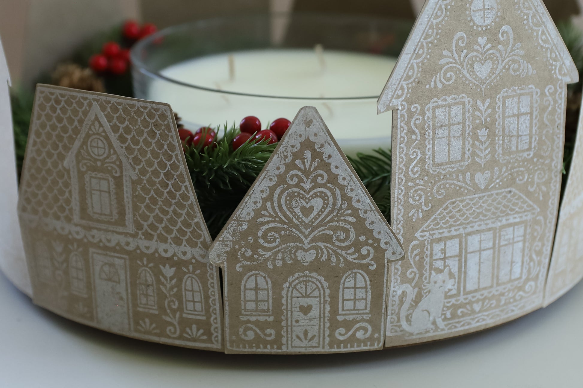 A close-up of a decorative holiday candle holder made from three gingerbread house cutouts in beige and white, surrounding a glass candle with white wax and red berries. Gingerbread Lane From Iron Orchid Designs.