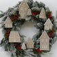 A festive Christmas wreath with frosted green pine branches, decorated with beige house ornaments, red berries, and small pinecones on a plain white background. Gingerbread Lane From Iron Orchid Designs.