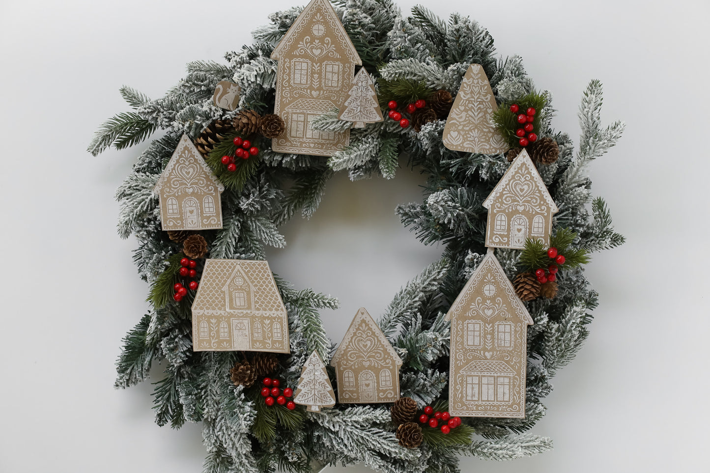 A festive Christmas wreath with frosted green pine branches, decorated with beige house ornaments, red berries, and small pinecones on a plain white background. Gingerbread Lane From Iron Orchid Designs.
