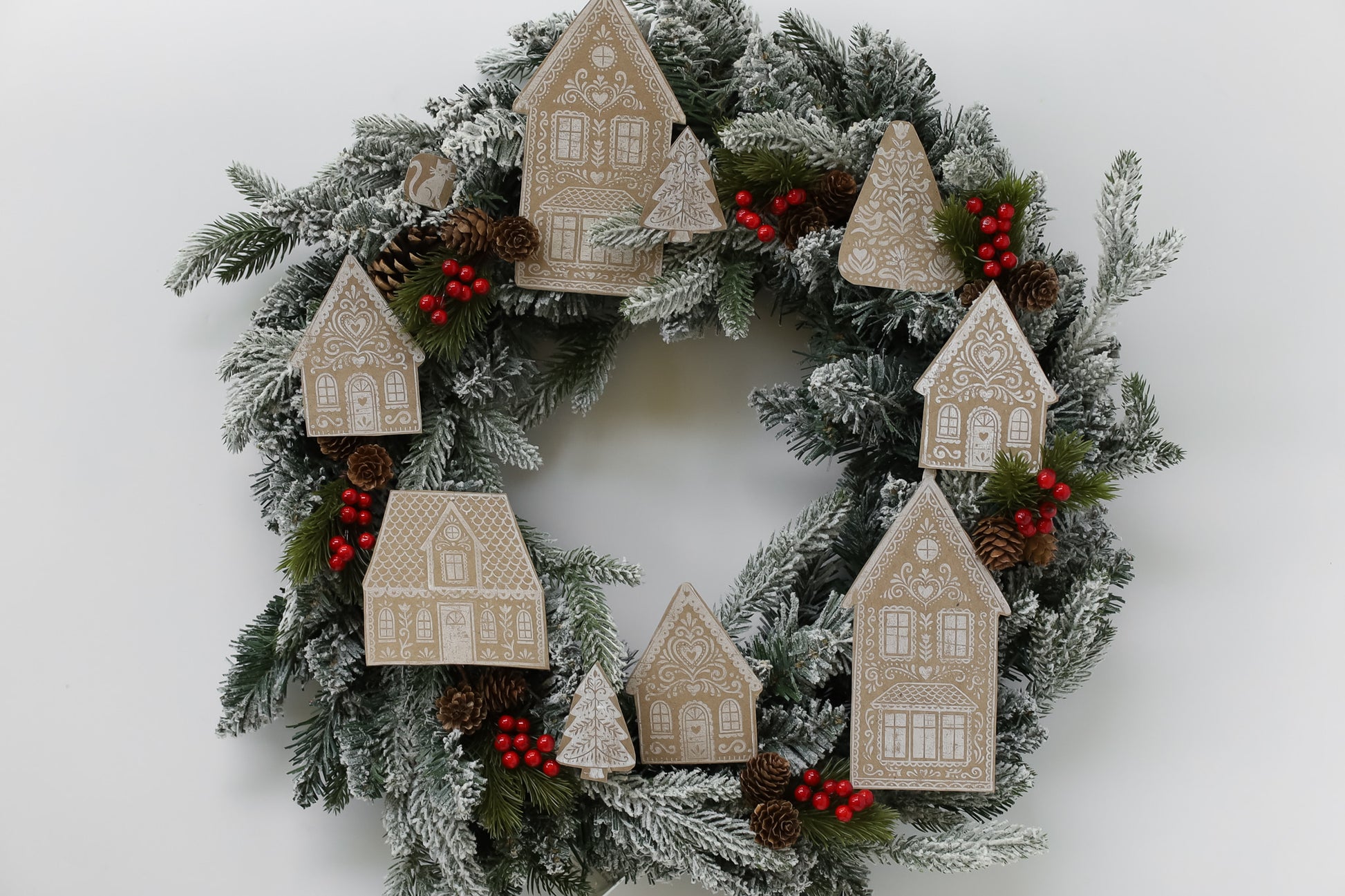 A festive Christmas wreath with frosted green pine branches, decorated with beige house ornaments, red berries, and small pinecones on a plain white background. Gingerbread Lane From Iron Orchid Designs.