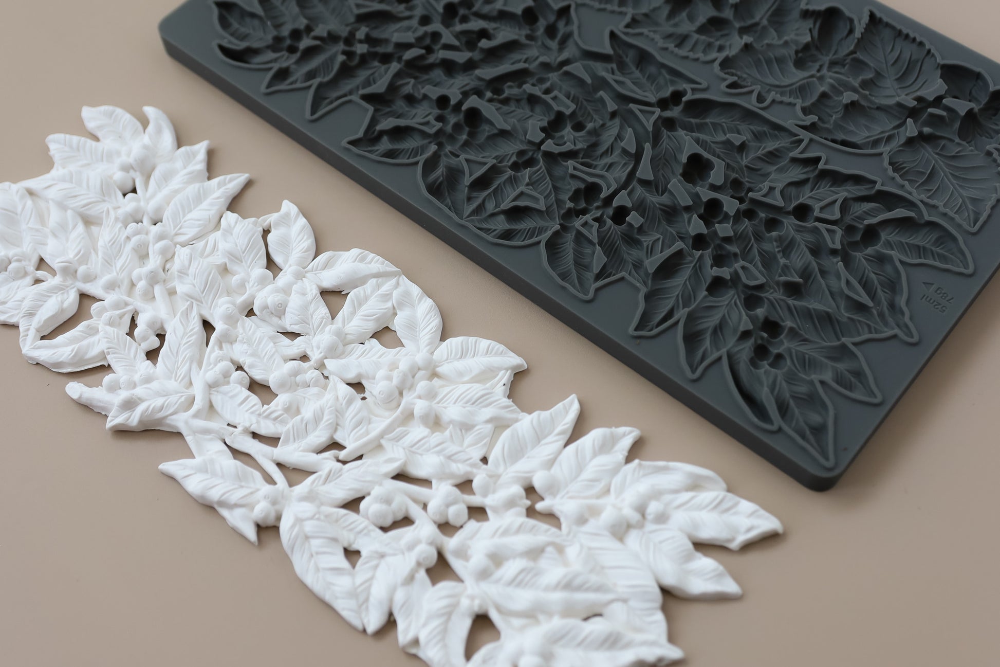White air dry clay leaf and berry floral trim shown next to a dark gray silicone mold that creates the same leaf pattern, on a solid beige background. Hazel and Bay mould. From Iron Orchid Designs.