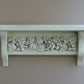 A white-painted wooden shelf with a decorative relief band of carved leaves and acorns, mounted on a plain taupe wall. From Iron Orchid Designs.