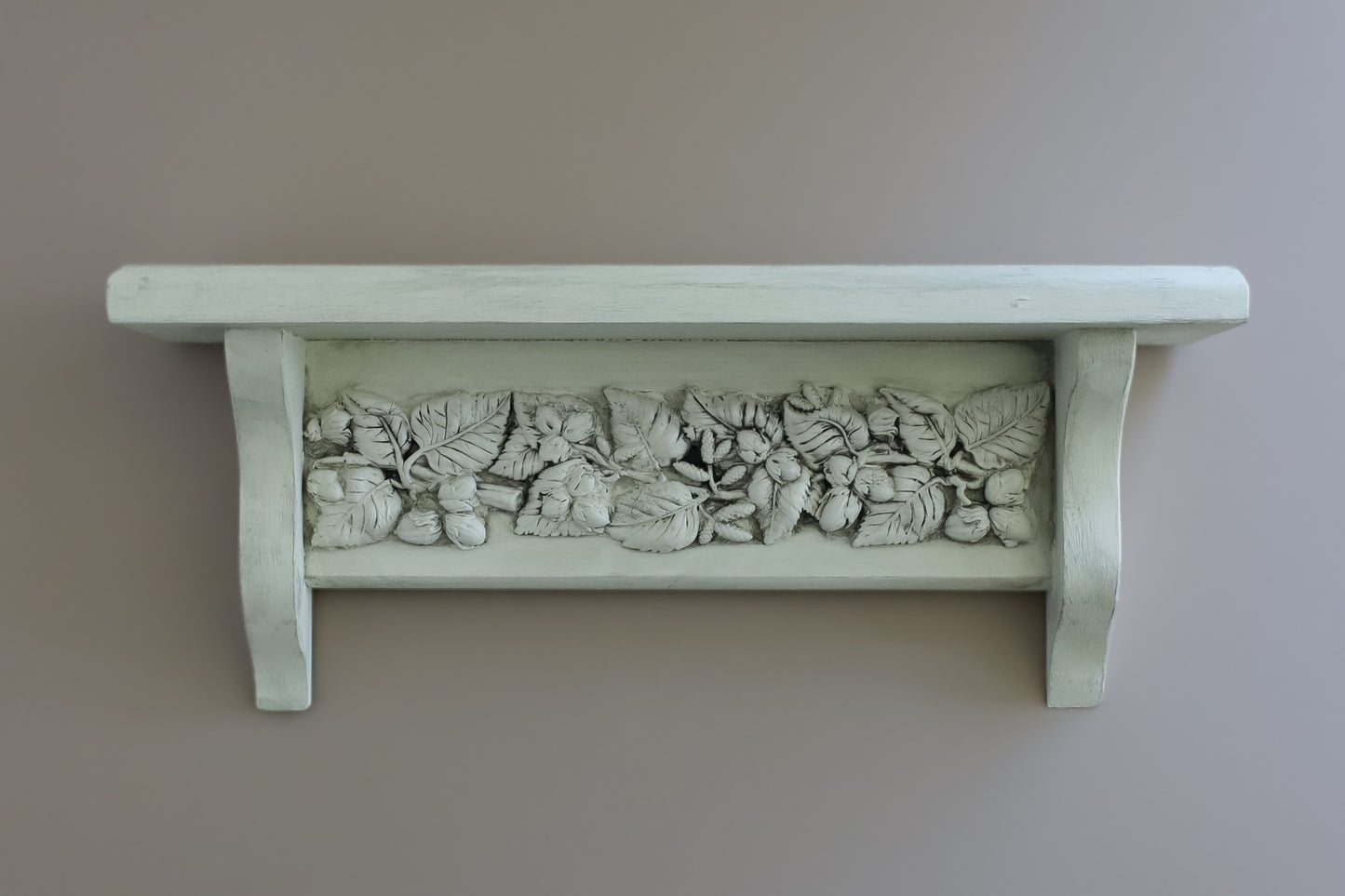 A white-painted wooden shelf with a decorative relief band of carved leaves and acorns, mounted on a plain taupe wall. From Iron Orchid Designs.