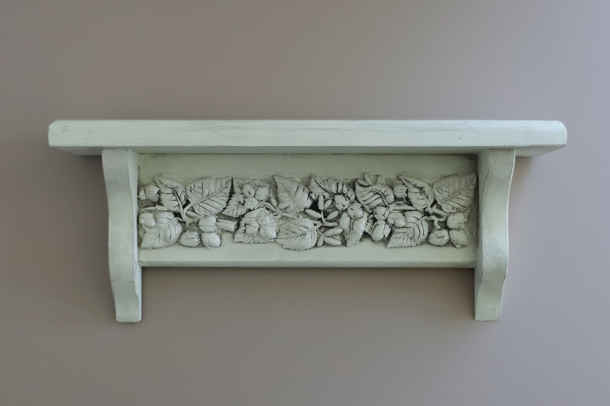 A white-painted wooden shelf with a decorative relief band of carved leaves and acorns, mounted on a plain taupe wall. From Iron Orchid Designs.