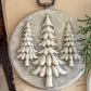 A round gray decorative plaque featuring three raised white pine tree silhouettes, styled on a beige surface with a pinecone and greenery nearby. Boughs of Holly Iron Orchid Designs