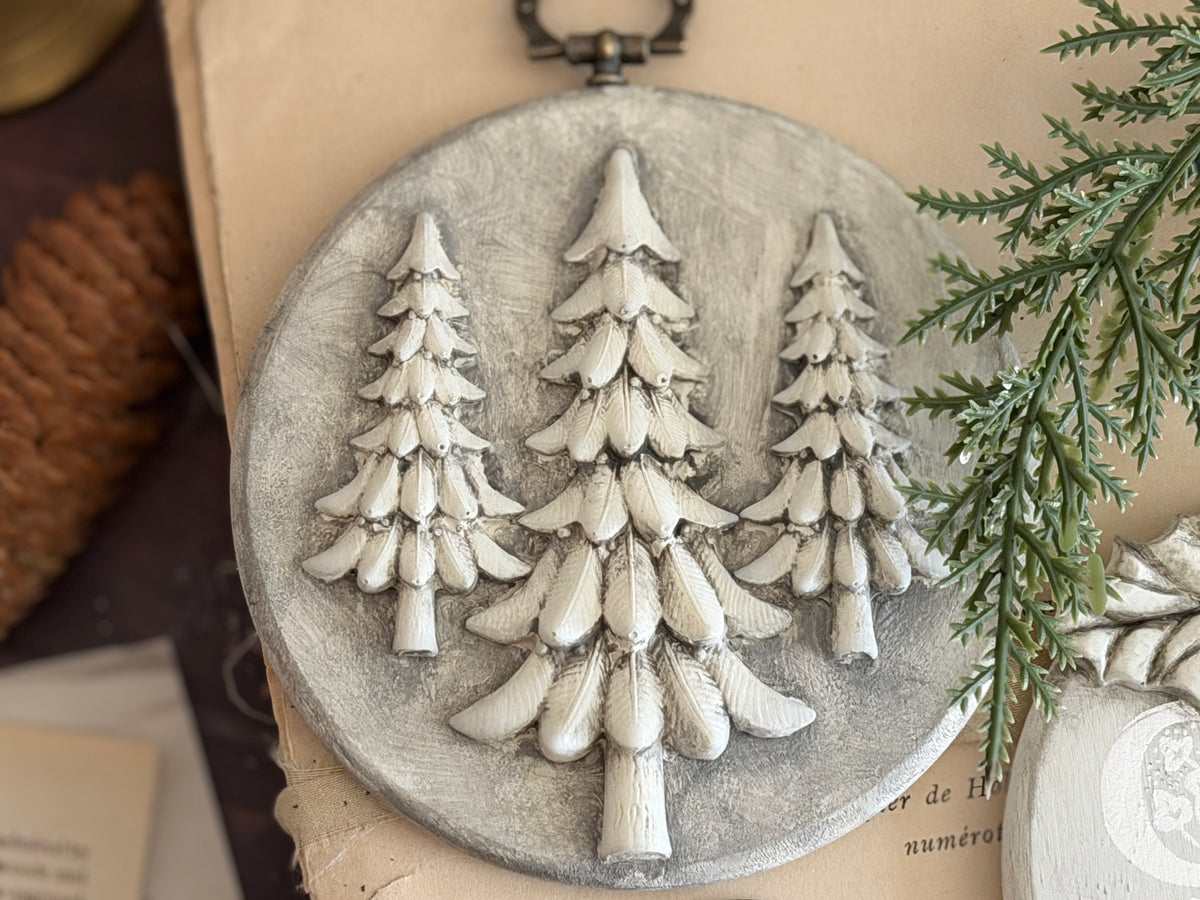 A round gray decorative plaque featuring three raised white pine tree silhouettes, styled on a beige surface with a pinecone and greenery nearby. Boughs of Holly Iron Orchid Designs