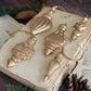 Five metallic gold Christmas ornament shapes arranged on an old open book with vintage paper pages, pinecones, and evergreen branches on a dark wooden surface. Baubles mould by Iron Orchid Designs.