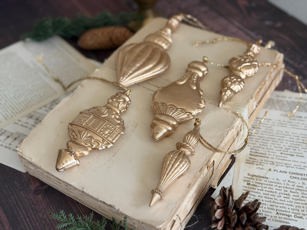 Five metallic gold Christmas ornament shapes arranged on an old open book with vintage paper pages, pinecones, and evergreen branches on a dark wooden surface. Baubles mould by Iron Orchid Designs.