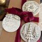 Three round Christmas ornament tags with raised holiday designs and text, tied to a kraft gift box with a dark red ribbon. The ornaments show a snowflake, pine trees, and the phrase 'Christmas in Art & Song' on a rustic tabletop with book pages and pinecones in the background. Boughs of Holly Iron Orchid Designs