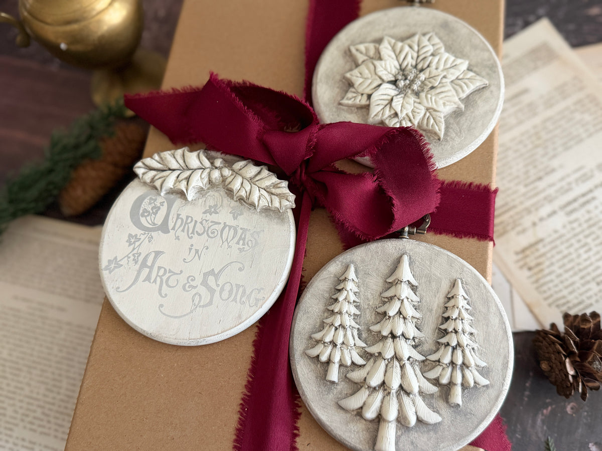 Three round Christmas ornament tags with raised holiday designs and text, tied to a kraft gift box with a dark red ribbon. The ornaments show a snowflake, pine trees, and the phrase 'Christmas in Art & Song' on a rustic tabletop with book pages and pinecones in the background. Boughs of Holly Iron Orchid Designs