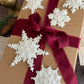 A brown kraft paper gift box wrapped with a deep burgundy ribbon and bow, decorated with five white lace snowflake ornaments, shown on a dark wooden surface. Snowflakes by Iron Orchid Designs