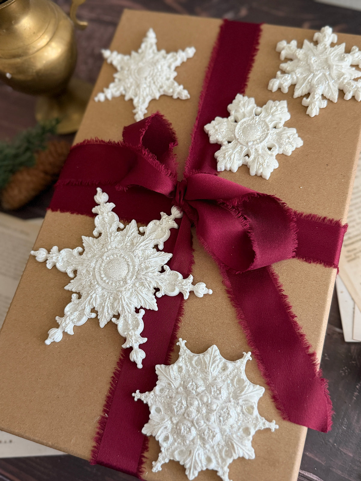 A brown kraft paper gift box wrapped with a deep burgundy ribbon and bow, decorated with five white lace snowflake ornaments, shown on a dark wooden surface. Snowflakes by Iron Orchid Designs