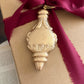A gold-toned decorative bottle-shaped ornament hanging from a dark magenta ribbon tied around a kraft paper gift box, with an open book visible in the background. Baubles mould by Iron Orchid Designs.