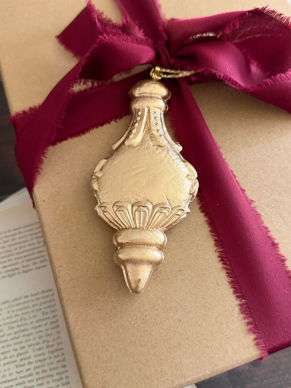 A gold-toned decorative bottle-shaped ornament hanging from a dark magenta ribbon tied around a kraft paper gift box, with an open book visible in the background. Baubles mould by Iron Orchid Designs.