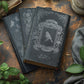 Decorative black books with ornate silver designs from Iron Orchid Designs Papillon transfer on a rustic wooden surface with green leaves.
