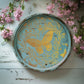 Decorative green wooden plate with Iron Orchid Designs Papillon butterfly design laying on a white surface with pink flowers