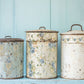 Three vintage-style metal canisters with floral patterns against a light blue wooden background